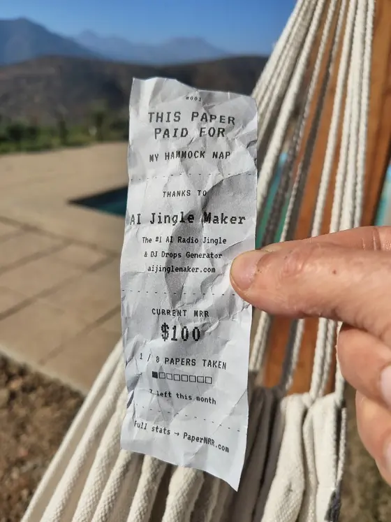 Hand holding a PaperMRR receipt that reads This Paper Paid For My Hammock Nap, sponsored by AI Jingle Maker, in front of a rope hammock with mountains in the background.
